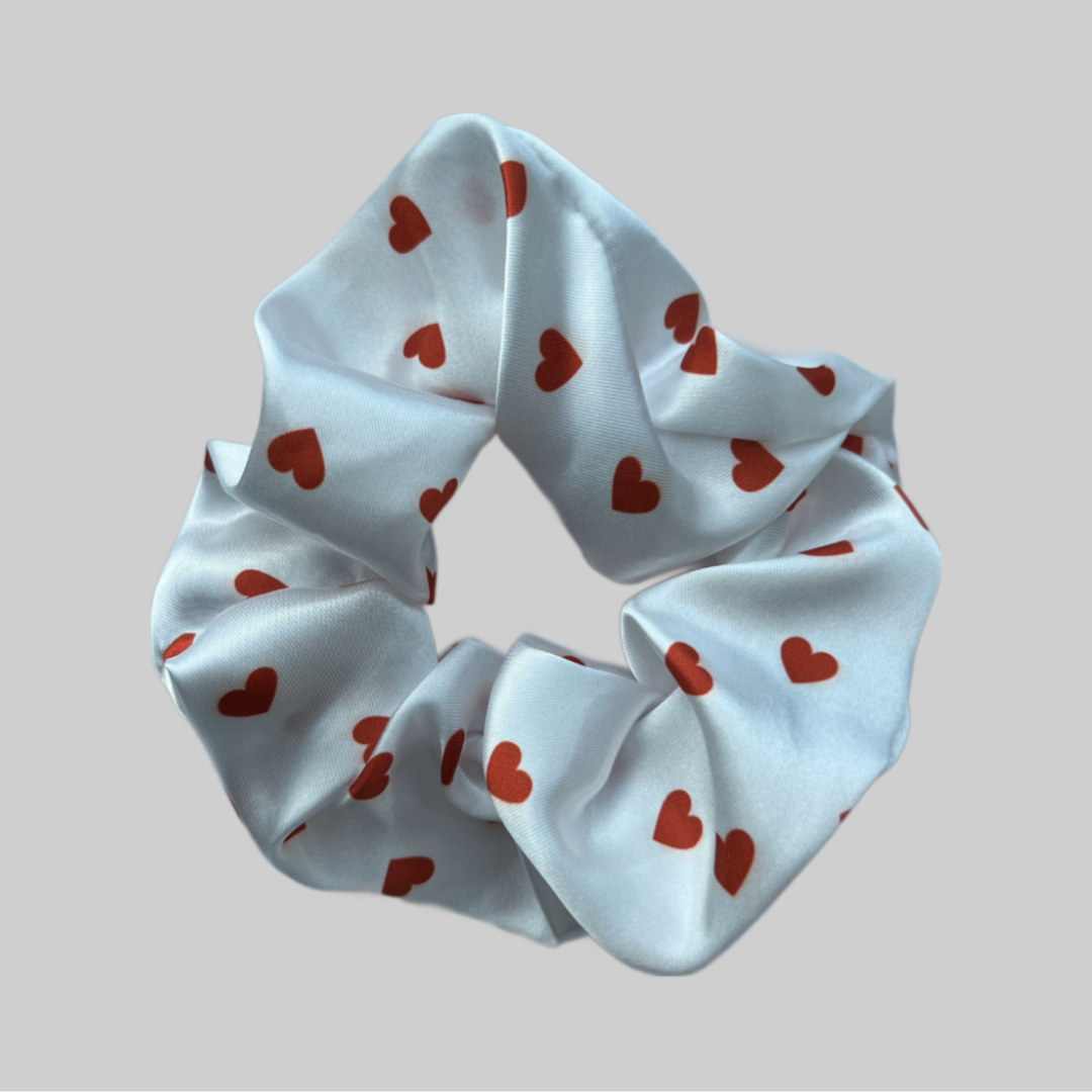 Flat lay of the Miae White Satin Scrunchie with red love hearts, a romantic and frizz-reducing hair accessory perfect for all hair types.